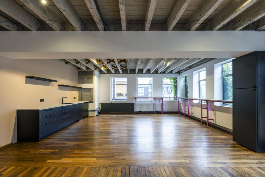 Ruime, lichtinvalrijke keuken en eetruimte aan de De Ruijterkade met houten vloer, zwart keukenblok en zichtbare plafondbalken.